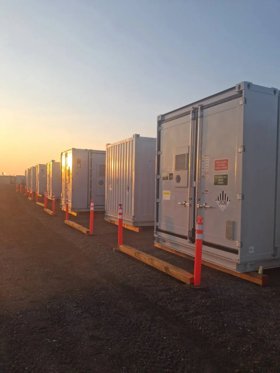 Row of UN3536-placarded battery energy storage containers resting on timber sleepers at sunset
