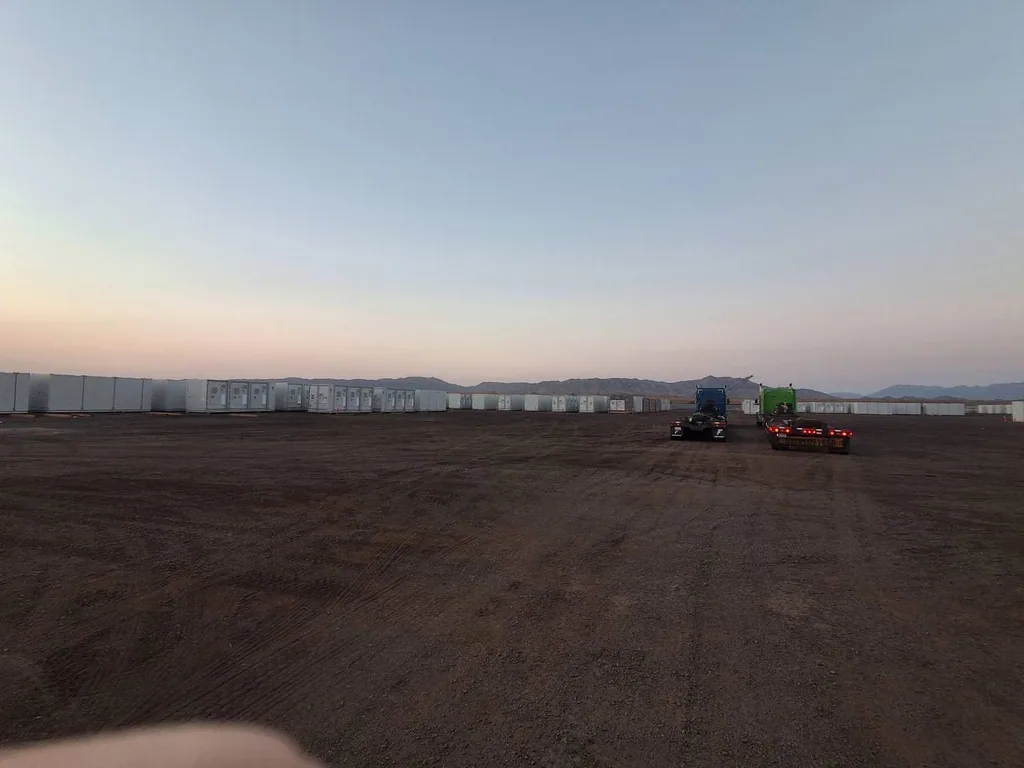 Utility-scale battery energy storage construction site at dusk with delivered containers and heavy-haul trucks on the horizon