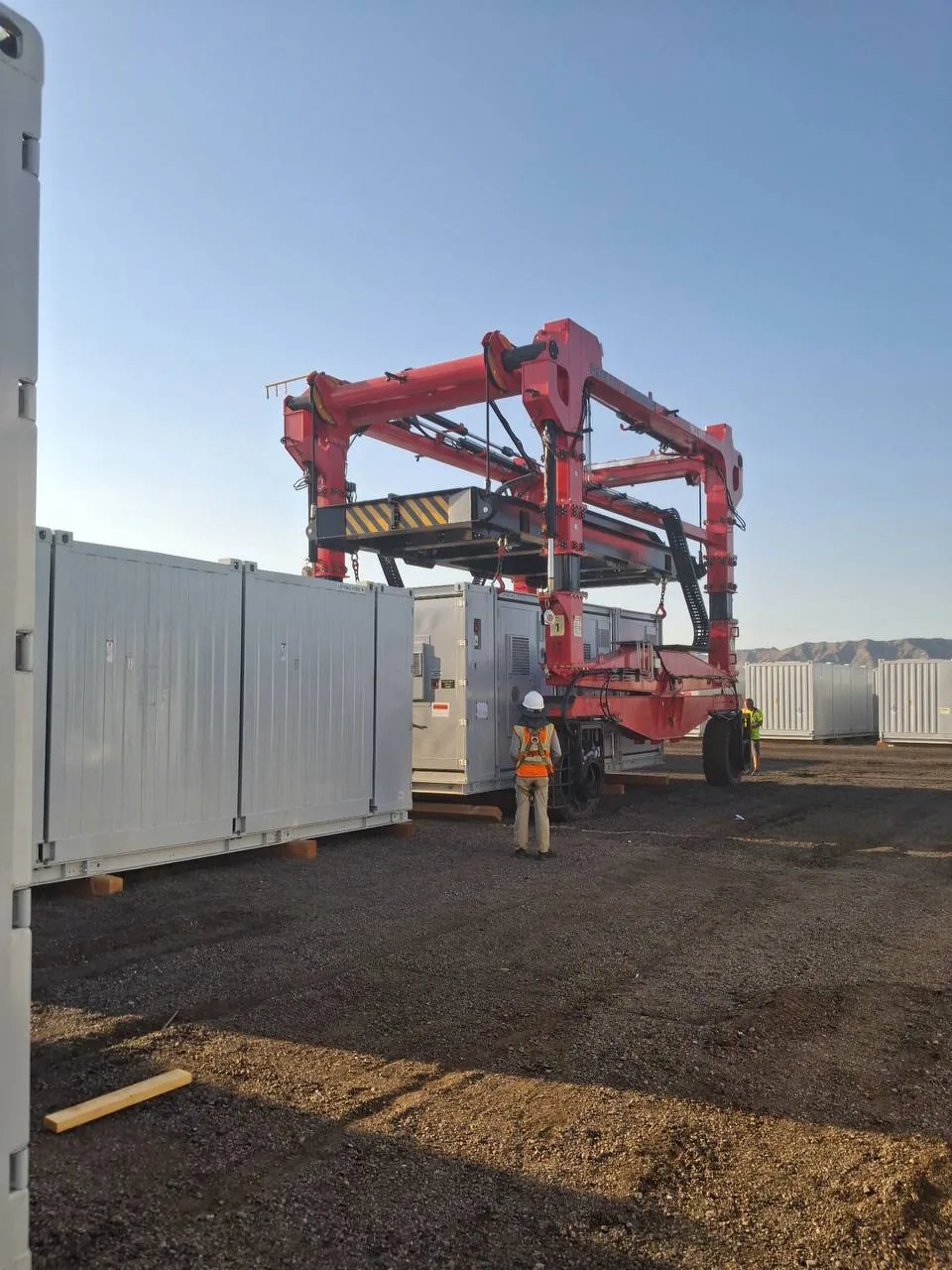 Red 60-tonne straddle carrier positioning a containerized battery energy storage unit while ground crew in high-visibility gear spot the lift